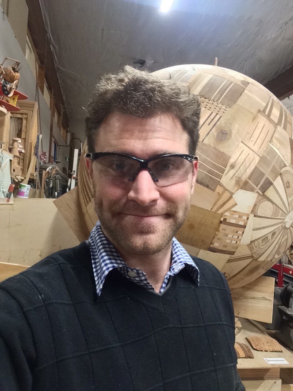 Selfie in safety glasses in a workshop, in front of a giant sphere made of patchwork wood