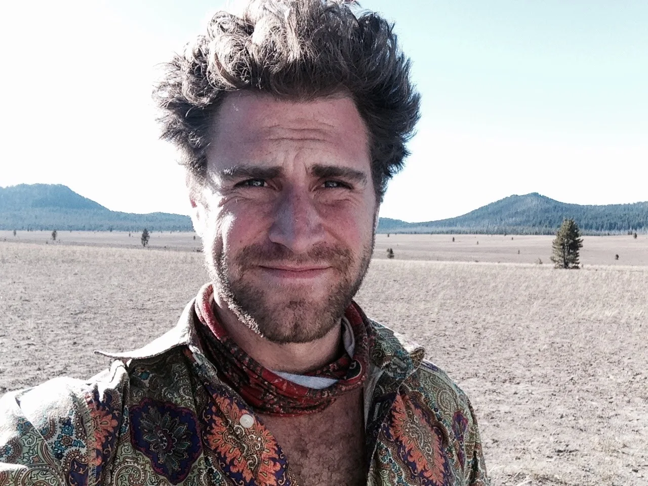 A windswept selfie in a paisley shirt and bandana on a wide high-desert plain, hills behind