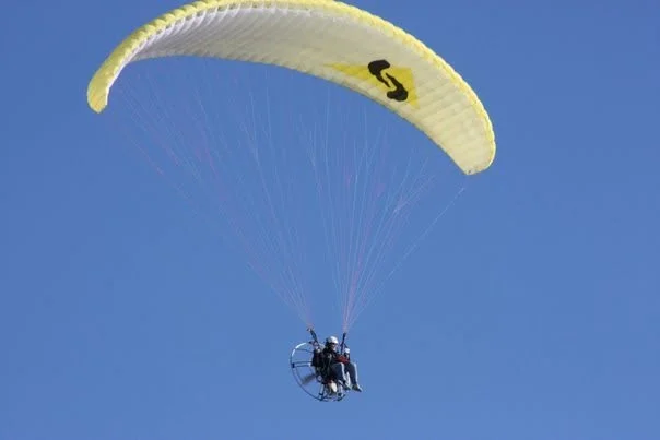 A paramotor under a yellow wing, high in a clear blue sky